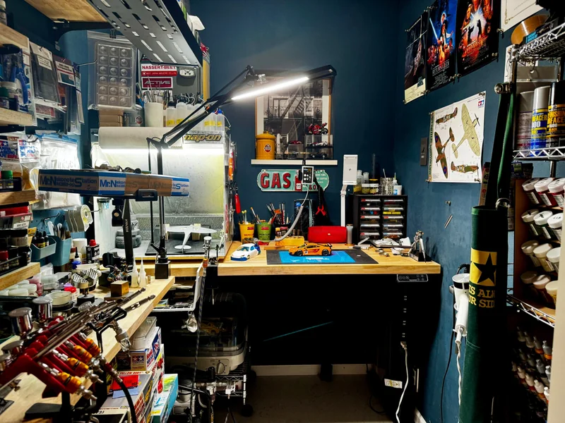 模型の作業部屋。塗装ブースとエアーブラシ、作業台の上の製作中のカーモデル、壁一面の塗料棚。